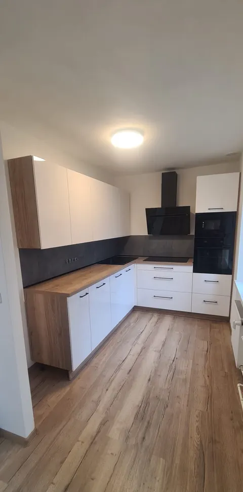 Kuchyňská linka do L, bílá lesklá dvířka, pracovní deska v dubovém dekoru, betonová zástěna, černá madla a černá digestoř