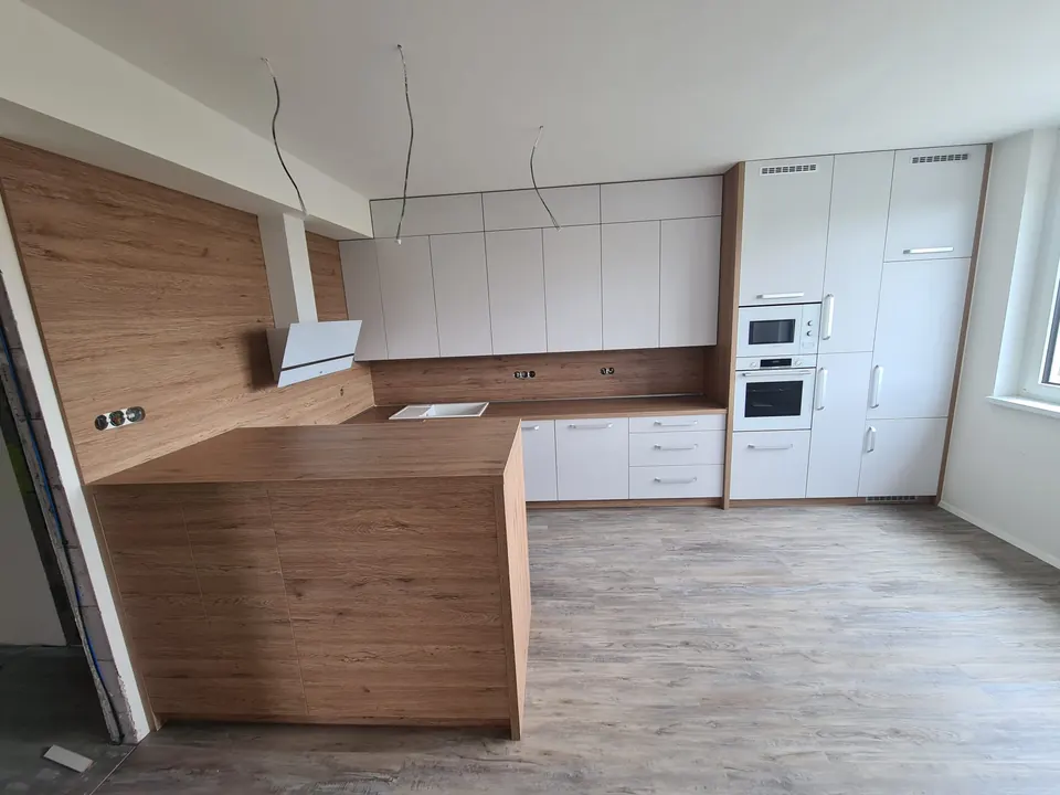 Kuchyňská linka do U s barovým pultem v dubovém dekoru, bílá matná dvířka s úchytkami, dubová pracovní deska se zástěnou a bílé vestavné spotřebiče