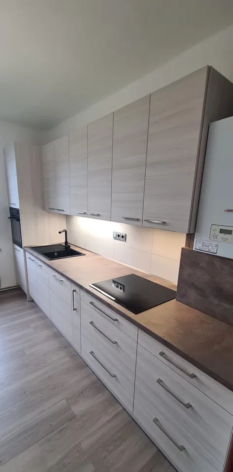 Kuchyňská linka podél stěny, dvířka ve světlém dřevěném dekoru, tmavá pracovní deska, černý dřez a černá varná deska