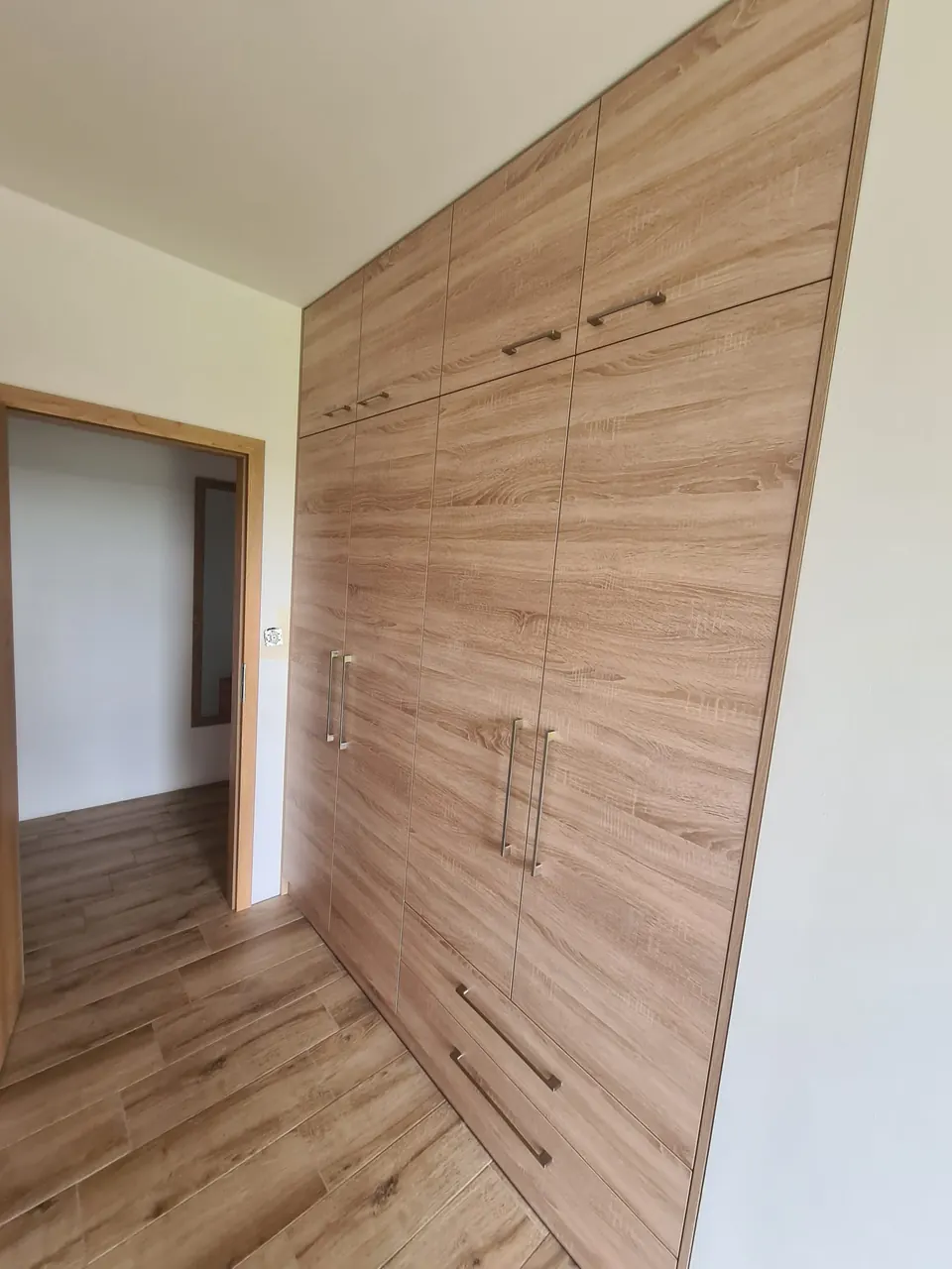 Vestavěná skříň přes celou stěnu v dubovém dekoru, dvířka bez rámu se stříbrnými úchytkami, nadstavec a spodní zásuvky